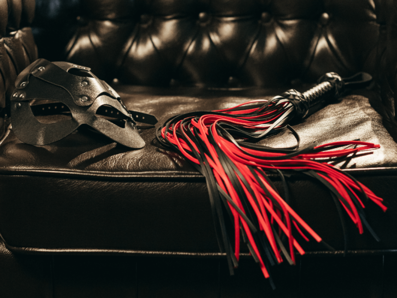 BDSM Mythen Symbolbild: rote Peitsche und Ledermaske auf Ledercouch BDSM myths Symbolic image: red whip and leather mask on leather couch