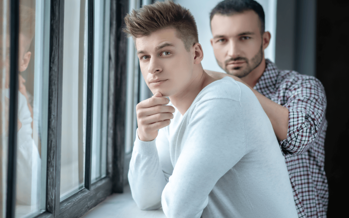 Vertrauensbruch in der Beziehung. Symbolbild: Zwei Männer stehen ernst dreinblickend an einem Fenster Breach of trust in a relationship. Symbolic image: Two men standing at a window looking serious