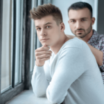 Vertrauensbruch in der Beziehung. Symbolbild: Zwei Männer stehen ernst dreinblickend an einem Fenster Breach of trust in a relationship. Symbolic image: Two men standing at a window looking serious