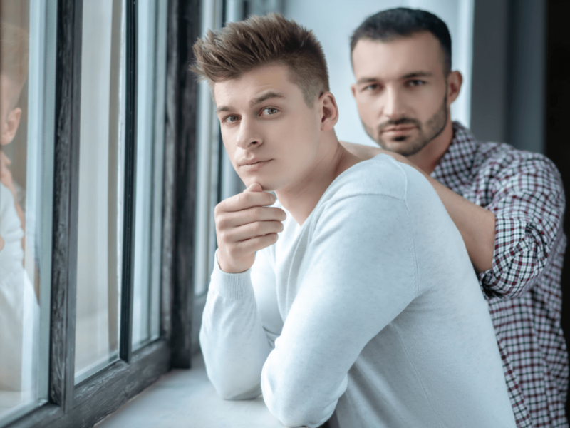 Vertrauensbruch in der Beziehung. Symbolbild: Zwei Männer stehen ernst dreinblickend an einem Fenster Breach of trust in a relationship. Symbolic image: Two men standing at a window looking serious