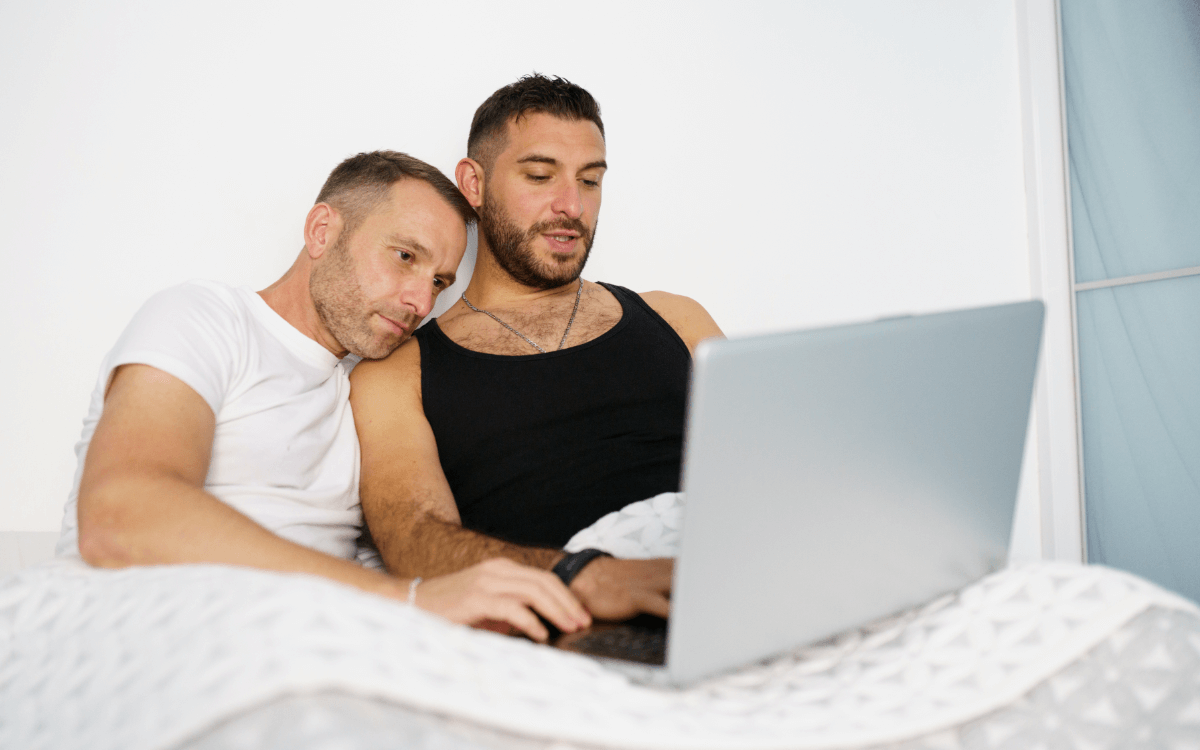 Beziehungsprobleme schwule Paare. Symbolbild: Zwei Männer liegen im Bett und schauen gemeinsam in den Laptop. Englisch: Relationship problems of gay couples. Symbolic image: Two men lying in bed and looking at a laptop together.