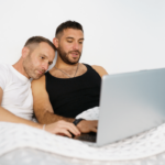 Beziehungsprobleme schwule Paare. Symbolbild: Zwei Männer liegen im Bett und schauen gemeinsam in den Laptop. Englisch: Relationship problems of gay couples. Symbolic image: Two men lying in bed and looking at a laptop together.