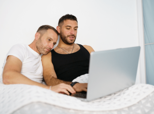 Beziehungsprobleme schwule Paare. Symbolbild: Zwei Männer liegen im Bett und schauen gemeinsam in den Laptop. Englisch: Relationship problems of gay couples. Symbolic image: Two men lying in bed and looking at a laptop together.