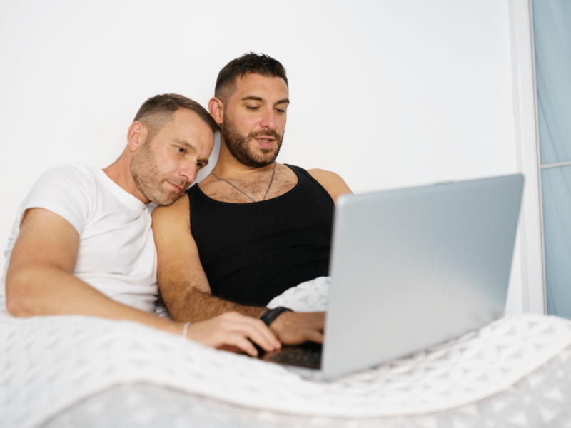 Beziehungsprobleme schwule Paare. Symbolbild: Zwei Männer liegen im Bett und schauen gemeinsam in den Laptop. Englisch: Relationship problems of gay couples. Symbolic image: Two men lying in bed and looking at a laptop together.