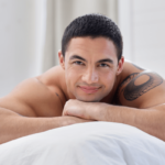 Prostata Massage Symbolbild: Ein Mann mit freiem Oberkörper liegt auf dem Bauch und lächelt in die Kamera. Englisch: Prostate massage, symbolic image: a naked man is lying on his front smiling at the camera.