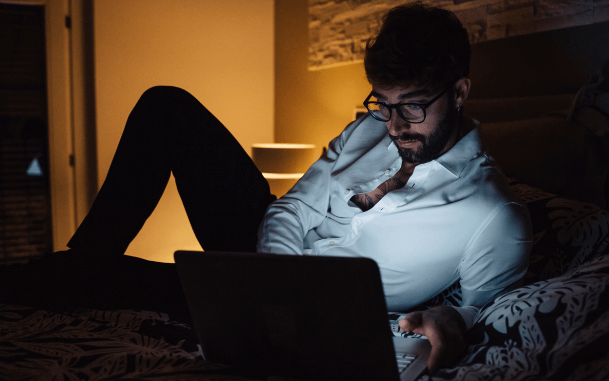 Pornosucht. Symbolbild: Ein Mann liegt im Halbdunkeln vor seinem Laptop auf dem Bett. Englisch: Porn addiction. Symbolic image: a man is lying on his bed in front of his laptop in semi-darkness.