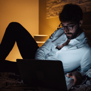 Pornosucht. Symbolbild: Ein Mann liegt im Halbdunkeln vor seinem Laptop auf dem Bett. Englisch: Porn addiction. Symbolic image: a man is lying on his bed in front of his laptop in semi-darkness.