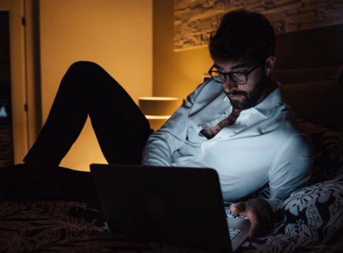 Pornosucht. Symbolbild: Ein Mann liegt im Halbdunkeln vor seinem Laptop auf dem Bett. Englisch: Porn addiction. Symbolic image: a man is lying on his bed in front of his laptop in semi-darkness.