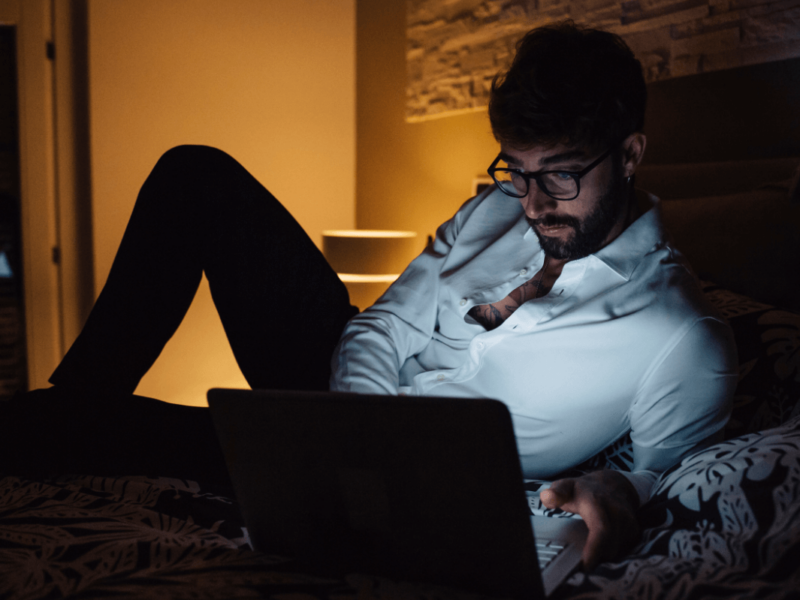 Pornosucht. Symbolbild: Ein Mann liegt im Halbdunkeln vor seinem Laptop auf dem Bett. Englisch: Porn addiction. Symbolic image: a man is lying on his bed in front of his laptop in semi-darkness.