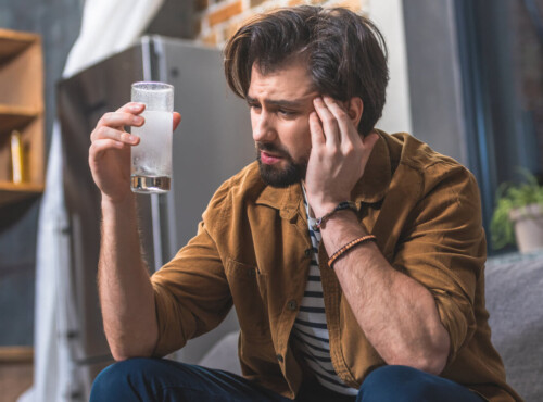 Poppers Nebenwirkungen Symbolbild: Ein Mann hält sich den Kopf vor Schmerzen und hält ein Glas mit Schmerzmittel in der Hand. Englisch: Side effects of poppers Symbolic image: A man is holding his head in pain and holding a glass of painkillers in his hand.