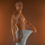 Penislänge Symbolbild: Ein Mann mit nacktem Oberkörper hält seine Hose im Schritt soweit von sich weg, dass ein langer Penis angedeutet wird. Englisch: Penis length Symbolic image: A man with a bare upper body holds his pants away from his crotch, suggesting a long penis.