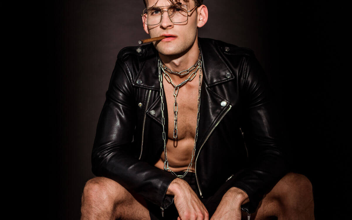 Fetisch vs. Kink Symbolbild: Ein Mann mit offener Lederjacke und Zigarre im Mund schaut lassziv in die Kamera. Fetish vs. Kink Symbolic image: A man with an open leather jacket and a cigar in his mouth looks lasciviously into the camera.