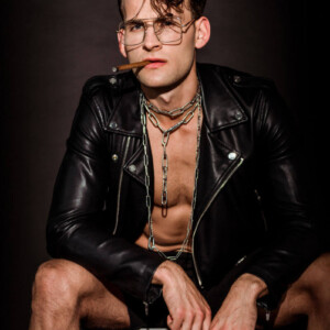 Fetisch vs. Kink Symbolbild: Ein Mann mit offener Lederjacke und Zigarre im Mund schaut lassziv in die Kamera. Fetish vs. Kink Symbolic image: A man with an open leather jacket and a cigar in his mouth looks lasciviously into the camera.